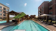 Located in Frisco, TX, this upscale outdoor amenity space centers around a generously sized, crystal-clear swimming pool with lap lanes, designed to evoke a resort-like atmosphere. The pool deck is finished with elegant brick pavers and lined with a generous arrangement of lounge chairs and umbrellas in vibrant green and neutral tones, offering ample space for sunbathing and relaxation. A dedicated spa provides an additional retreat adjacent to the main pool.

A standout feature of the space is the covered cabana, constructed with rustic wood beam framing and a pitched roof supported by brick columns. Beneath the structure, comfortable wicker lounge chairs, accent tables, and plush seating create a shaded gathering area ideal for socializing or unwinding poolside. Unique driftwood accent pieces add a natural, artistic touch to the setting.

Outdoor dining areas with tables and chairs are thoughtfully positioned throughout the courtyard, while gas grills provide convenient amenities for outdoor cooking. The entire pool area is enclosed by a wrought-iron fence, ensuring privacy and security.

The space is framed on multiple sides by multi-story brick residential buildings featuring private balconies, arched entryways, and large windows — an architectural design that lends a sense of community and upscale urban living. Abundant natural light floods the courtyard under the open Texas sky.