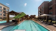 Located in Frisco, TX, this upscale outdoor amenity space centers around a generously sized, crystal-clear swimming pool with lap lanes, designed to evoke a resort-like atmosphere. The pool deck is finished with elegant brick pavers and lined with a generous arrangement of lounge chairs and umbrellas in vibrant green and neutral tones, offering ample space for sunbathing and relaxation. A dedicated spa provides an additional retreat adjacent to the main pool.

A standout feature of the space is the covered cabana, constructed with rustic wood beam framing and a pitched roof supported by brick columns. Beneath the structure, comfortable wicker lounge chairs, accent tables, and plush seating create a shaded gathering area ideal for socializing or unwinding poolside. Unique driftwood accent pieces add a natural, artistic touch to the setting.

Outdoor dining areas with tables and chairs are thoughtfully positioned throughout the courtyard, while gas grills provide convenient amenities for outdoor cooking. The entire pool area is enclosed by a wrought-iron fence, ensuring privacy and security.

The space is framed on multiple sides by multi-story brick residential buildings featuring private balconies, arched entryways, and large windows — an architectural design that lends a sense of community and upscale urban living. Abundant natural light floods the courtyard under the open Texas sky.