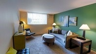 This single-floor space centers around a thoughtfully designed living room that balances comfort and contemporary style. The room is defined by a striking teal accent wall, which serves as a bold focal point and provides a vivid contrast against the warm, neutral-toned surrounding walls. Two abstract artworks hang on the teal wall, adding a curated, gallery-like quality to the space.

The living area is furnished with a curved gray sofa paired with a rounded upholstered ottoman, offering a cozy and inviting seating arrangement. A sleek, modern armchair with a chrome frame and leather upholstery complements the contemporary aesthetic, while a classic floor lamp provides warm ambient lighting. The carpeted flooring adds texture and softness underfoot, contributing to the overall comfort of the space.

A large window fitted with vertical blinds allows generous natural light to fill the room while offering a view of outdoor greenery. A wall-mounted air conditioning unit beneath the window provides climate control. The overall ambiance is modern and welcoming, with a carefully considered mix of color, texture, and furnishings.