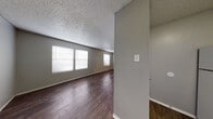 This single-floor unit offers a straightforward, functional layout with a cohesive neutral aesthetic carried throughout each room. Dark hardwood-style flooring runs continuously across the living areas and bedroom, providing a warm contrast against the light gray walls and white baseboards. Textured popcorn ceilings are present throughout the unit, adding a classic finishing detail overhead.

The main living area is open and well-proportioned, featuring multiple windows fitted with horizontal blinds that allow natural light to filter in while maintaining privacy. The space flows naturally from the entry door, which opens directly into the living area, making the layout feel accessible and easy to navigate.

The kitchen is compact and efficiently arranged, equipped with white cabinetry, a gray laminate countertop, a built-in dishwasher, a full-size refrigerator, and a single-basin sink. Upper cabinets provide additional storage above the counter space.

The bedroom is a quiet, enclosed room with a window that brings in natural light. The bathroom includes a vanity with a gray countertop, an undermount sink, and a large mirror that adds a sense of depth to the space. The overall unit presents a clean, move-in-ready environment with consistent finishes throughout.