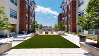 This community space features two distinct areas designed to support an active lifestyle. The open-air courtyard is framed by multi-story brick buildings and showcases a manicured turf lawn at its center, surrounded by wide concrete pavers. Modular wood-framed seating clusters and planters with young trees provide comfortable gathering spots, while lounge chairs offer additional relaxation options. The courtyard's open layout invites ample natural light and provides views of the surrounding urban skyline.

The fitness center is a comprehensively equipped facility spread across a single floor. Herringbone-patterned carpet tiles cover the floor throughout, adding a modern aesthetic. Large windows along multiple walls flood the space with natural light and offer outdoor views, while ceiling fans and recessed lighting ensure comfort during workouts. Full-length mirrors line portions of the walls, visually expanding the room.

The gym is stocked with a wide variety of equipment, including treadmills, a rowing machine, a stationary cycle, a multi-functional cable and power rack system, a dumbbell rack, medicine balls, stability balls, battle ropes, and a cable-based strength machine. A dedicated cardio zone with a wall-mounted television is positioned near the windows, creating an bright and motivating environment. Built-in shelving along one wall provides additional storage and organization for accessories.