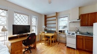 Located in Cambridge, MA, this single-floor unit offers a well-organized and functional layout that makes efficient use of its space. Throughout the unit, warm honey-toned hardwood floors provide a consistent and inviting foundation.

The bedroom is a bright, comfortable space illuminated by a pair of large diamond-pane windows that allow natural light to fill the room. It includes ample space for a bed and a dedicated desk area, with a dresser providing additional storage.

The kitchen is efficiently laid out with warm wood cabinetry, a granite-style countertop, a full-size refrigerator, and a white electric range with a ventilation hood above. White tile backsplash adds a clean, classic touch to the cooking area. A sliding wood-panel door provides a practical partition nearby.

Adjacent to the kitchen, the open living and dining area is framed by two diamond-pane windows, flooding the space with natural light. A built-in wooden open shelving unit anchors the area, while a compact dining table with chairs and a desk workspace are neatly arranged to maximize utility.

The bathroom is finished with light tile flooring and tiled walls, featuring a vanity with storage, a toilet, and a bathtub with a sliding glass enclosure. Overall, this Cambridge unit presents a practical and comfortable living environment with classic architectural details.