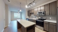 This well-appointed single-floor residence showcases a thoughtfully designed open-concept layout that seamlessly connects the kitchen and living areas. The kitchen is a standout feature, offering rich taupe cabinetry with sleek hardware, a subway tile backsplash, white quartz countertops, and a full suite of stainless steel appliances including a refrigerator, electric range, microwave, and dishwasher. A central island with a matching quartz surface and pendant lighting above provides additional prep space and casual seating. Track lighting illuminates the kitchen, while a ceiling fan anchors the adjacent living area, which benefits from natural light streaming through a large window with horizontal blinds. Light wood-look flooring flows throughout the main living and kitchen areas, adding warmth and continuity. The bathroom is efficiently designed with a combination tub and shower, large-format tile flooring, a framed mirror, and a dark-cabinetry vanity topped with a white surface and dual fixtures. The bedroom is a comfortable retreat, featuring plush carpeting, a ceiling fan, and a generously sized window that fills the room with natural light. The overall aesthetic is clean, contemporary, and move-in ready.