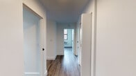 This single-floor unit showcases a clean, modern design highlighted by wood-look flooring that runs consistently throughout the living areas and bedrooms. A central hallway connects the various rooms, providing an efficient and organized layout visible in the 3D floor plan.

Each bedroom features crisp white walls, large windows with black frames that frame urban cityscape views, and individual wall-mounted PTAC heating and cooling units. Natural light flows generously into the rooms, enhancing the bright, airy atmosphere.

The kitchen is outfitted with light gray shaker-style cabinetry featuring black hardware, a white hexagon tile backsplash, light-toned countertops, a single-basin stainless steel sink with a matte black faucet, and stainless steel appliances including an over-the-range microwave.

The bathroom is a standout space, featuring contrasting large-format dark hexagon floor tiles paired with white hexagon mosaic wall tiles extending into the tub surround. A gray vanity with black hardware, a mirrored medicine cabinet, recessed lighting, and matte black fixtures complete the refined, contemporary aesthetic throughout.