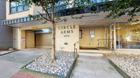 Circle Arms, located at 2416, is a well-established residential building characterized by its mid-century architectural style. The exterior presents a clean yellow brick façade with understated signage, a ground-level parking garage with roll-up access, and a welcoming entry with low-profile steps and iron railings accented by a landscaped planter featuring river rock and a mature shade tree. The building rises several stories, reflecting the structured, utilitarian design sensibility of its era.

The single-floor unit interior is finished in crisp white throughout, lending a clean and neutral aesthetic. Interior doors feature classic brass knob hardware, maintaining a traditionally styled appearance. A notable feature of the unit is its dedicated storage closet, which includes multiple painted wood shelving units arranged along the walls, offering practical and well-organized storage capacity across several tiers.

The surrounding urban environment, visible from the building's exterior vantage point, includes mature deciduous trees and neighboring multi-story buildings, providing a lively city context. The building's integration of parking at street level adds a practical convenience for residents. Overall, Circle Arms presents a functional, no-frills residential space suited to urban living.