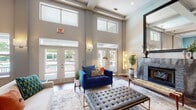 Located at 8531 Summersweet Ln in Raleigh, NC, this thoughtfully designed single-floor space offers an impressive blend of elegance and functionality. The main lounge area commands attention with dramatically soaring ceilings that rise to reveal clerestory windows, flooding the interior with abundant natural light. Warm hardwood flooring flows throughout, anchoring a sophisticated seating arrangement that includes plush velvet accent chairs and a tufted leather bench centered around a striking fireplace with a bold tile surround and an oversized mirror above the mantle. French doors open to an outdoor area, further enhancing the sense of openness and connection to the surrounding greenery. A wide, airy corridor connects the lounge to the rest of the space, showcasing vibrant botanical artwork and additional seating vignettes set against tall, neutral-toned walls. The kitchen is a standout feature, distinguished by vivid teal accent walls and soaring ceilings that carry the sense of volume throughout. Rich wood cabinetry pairs with granite countertops, stainless steel appliances, and a spacious island, creating a practical yet visually dynamic culinary environment. Together, these spaces form a cohesive, design-forward interior that balances comfort with contemporary style.
