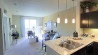 This single-floor space showcases an open-concept layout that seamlessly connects the kitchen, dining, and living areas into one cohesive environment. The kitchen features a granite-style countertop with a double stainless steel sink and a classic chrome faucet. Three elegant pendant lights hang above the counter, providing warm, focused illumination that complements the overall brightness of the space. The dining area sits adjacent to the kitchen, furnished with a sleek glass table and coordinating chairs, creating a modern and airy feel. The living area is carpeted and comfortably arranged, featuring a patterned accent chair, a low-profile sofa, and a round coffee table, giving the space a relaxed yet polished character. Neutral, soft green-toned walls contribute to a calm and welcoming ambiance throughout. A large sliding glass door floods the interior with abundant natural light and opens to a balcony with open views beyond, enhancing the sense of spaciousness. The cohesive design choices, from lighting to furnishings, create a functional and inviting single-level living environment.