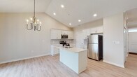 This single-floor home offers a bright and airy open-concept layout anchored by a modern kitchen. The kitchen is outfitted with crisp white cabinetry, stainless steel appliances including a refrigerator, range, and microwave, a center island with a sink and light countertops, and a tile backsplash. Recessed lighting and a stylish chandelier above the dining area add warmth to the space. Light wood-look flooring flows throughout the main living areas, complementing the neutral warm-toned walls.

The living room impresses with soaring vaulted ceilings and a ceiling fan, creating a generous sense of height and openness. A window allows natural light to brighten the room. A hallway leads to the private quarters, including a carpeted bedroom with crown molding, a ceiling fan, and a window with blinds. The bedroom is served by a spacious walk-in closet with wire shelving. The primary bathroom features a dual-sink vanity with white cabinetry and quartz-style countertops, a glass-enclosed shower, and wood-look flooring. A secondary half bath offers a single vanity and toilet. The home also includes an attached garage with a concrete floor and overhead door mechanism.