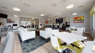Located at 146 Chesnut Crossing Road in Newark, Delaware, this single-story clubhouse presents a contemporary, open-concept design that seamlessly integrates multiple functional zones within one expansive space. Light hardwood-style flooring runs continuously throughout, lending visual cohesion and a clean, modern aesthetic.

The fully equipped kitchen features stainless steel appliances, including a full-size refrigerator, wall ovens, and a dishwasher. Dark wood cabinetry with chrome hardware is paired with white quartz countertops, creating a striking two-tone contrast. Pendant lighting and a wall-mounted flat-screen television enhance its functionality and style.

The lounge area is anchored by a bold geometric-patterned area rug and furnished with crisp white sofas and a colorful upholstered ottoman. Pendant lights hang overhead, while vibrant artwork accents the walls. Adjacent to the lounge, high-top tables with sleek gray upholstered chairs are positioned along a wall of expansive windows, flooding the space with natural light and offering views of the surrounding community.

A dining area with white tables and chartreuse seating adds a lively pop of color, while a glass-partitioned room visible in the background suggests additional workspace or meeting accommodations. The overall ambiance is bright, modern, and welcoming.