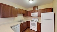 This single-floor residence offers a well-organized layout designed for comfortable everyday living. The kitchen is a functional centerpiece, featuring warm medium-toned wood cabinetry that provides generous storage space both above and below the counters. Light-colored countertops offer ample workspace, while the full suite of white appliances — including an electric range, over-the-range microwave, refrigerator, and dishwasher — ensures convenience. Wood-look flooring grounds the kitchen with a warm, durable finish. Adjacent to the kitchen, an open living and dining area is finished with plush neutral carpet and accented by a decorative chandelier, creating a welcoming gathering space. The layout flows naturally toward the private areas of the residence. The bedrooms feature the same soft carpeting, warm yellow-toned walls, and white trim, with windows fitted with horizontal blinds that allow natural light while maintaining privacy. Six-panel white doors add a classic touch throughout. The bathroom is clean and efficiently designed, with a wood-toned vanity, a wall-mounted medicine cabinet, bright vanity lighting, tile flooring, and a glass-enclosed walk-in shower. Overall, the space presents a cohesive, practical design with consistent warm tones and quality finishes throughout.