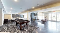 This single-floor amenity space is designed with a modern, open-concept layout that seamlessly blends recreation and relaxation. The interior is anchored by a substantial pool table set atop a bold geometric-patterned area rug, making it an immediate focal point upon entry. Polished concrete floors extend throughout, reflecting the recessed lighting above and contributing to the space's sleek, contemporary aesthetic.

A dedicated lounge area near the large windows features a linear fireplace framed in dark tile, surrounded by accent chairs and a sectional sofa arranged on a coordinating area rug. Natural light filters in through roller-shaded windows, creating a bright yet controlled atmosphere. The fireplace serves as a design centerpiece, adding warmth to the otherwise modern palette.

Toward the rear, a bar and café-style seating area offers counter-height stools and cabinetry finished in dark tones, ideal for casual gatherings or work sessions. Structural columns dressed in dark paneling add architectural definition, visually separating activity zones while maintaining an open, flowing feel. The overall color palette—neutral grays, warm taupes, and deep charcoals—creates a cohesive, sophisticated ambiance throughout.
