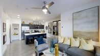This single-floor residence showcases a thoughtfully designed open-concept layout that seamlessly connects the kitchen and living areas into one harmonious living space. The kitchen is a standout feature, outfitted with dark navy cabinetry, stainless steel appliances including a French door refrigerator and built-in microwave, and a spacious island with a striking dark countertop and bar seating. Pendant lighting above the island adds a refined touch, complementing the recessed lighting throughout. The living area flows naturally from the kitchen and is bathed in a bright, airy atmosphere created by white walls and light wood-tone flooring. A modern ceiling fan anchors the space, providing both function and style. The living room is accented with a comfortable gray sofa adorned with yellow and blue decorative pillows, a coordinating accent chair, and a round white coffee table, all working together to create a warm yet contemporary feel. A large mirror leans against the wall, amplifying the sense of openness. Glass-paned interior doors offer a glimpse into an additional room beyond, adding architectural interest and transparency to the overall design. The cohesive color palette of navy, yellow, and gray ties every element of this well-appointed space together.