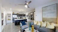 This single-floor residence showcases a thoughtfully designed open-concept layout that seamlessly connects the kitchen and living areas into one harmonious living space. The kitchen is a standout feature, outfitted with dark navy cabinetry, stainless steel appliances including a French door refrigerator and built-in microwave, and a spacious island with a striking dark countertop and bar seating. Pendant lighting above the island adds a refined touch, complementing the recessed lighting throughout. The living area flows naturally from the kitchen and is bathed in a bright, airy atmosphere created by white walls and light wood-tone flooring. A modern ceiling fan anchors the space, providing both function and style. The living room is accented with a comfortable gray sofa adorned with yellow and blue decorative pillows, a coordinating accent chair, and a round white coffee table, all working together to create a warm yet contemporary feel. A large mirror leans against the wall, amplifying the sense of openness. Glass-paned interior doors offer a glimpse into an additional room beyond, adding architectural interest and transparency to the overall design. The cohesive color palette of navy, yellow, and gray ties every element of this well-appointed space together.