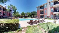 Located in Lewisville, Texas, this outdoor amenity space centers around a generously sized, resort-style swimming pool with strikingly clear, blue water. A raised spa is integrated directly into the pool deck, providing a seamless transition between the two water features and serving as a standout focal point of the area. The surrounding pool deck is finished in a light-toned concrete surface and is outfitted with multiple wood-framed chaise lounges, offering ample space for sunbathing and relaxation.

The complex features multi-story brick buildings with neutral stucco accents, private balconies, and large windows that overlook the pool courtyard, creating a sense of enclosure and community. A covered pavilion or cabana structure is visible along the perimeter, providing a shaded gathering area for residents. Ornamental iron fencing defines the boundary of the pool area, complemented by well-maintained landscaping including mature trees, trimmed hedges, and manicured greenery that add a lush, park-like atmosphere. The overall setting benefits from abundant natural Texas sunlight, contributing to an inviting, resort-inspired outdoor environment.