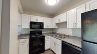 This two-story residence is designed with functionality and comfort in mind. The kitchen is well-appointed with crisp white shaker-style cabinetry accented by brushed nickel hardware, contrasting black appliances including an electric range, over-the-range microwave, and dishwasher. Gray laminate countertops and a double stainless steel sink complete the space, while durable wood-look vinyl flooring adds a clean, modern touch.

The open living area flows naturally from the kitchen, featuring the same warm wood-look flooring and neutral wall tones that lend a cohesive, inviting ambiance throughout the main level. A staircase connects the two floors, with the upper level housing the bedrooms.

The bedrooms are carpeted in soft gray tones and feature ceiling fans with integrated lighting. Each room benefits from natural light provided by windows fitted with horizontal blinds. Closet access is also present in the bedrooms, offering practical storage solutions.

The bathrooms maintain a clean, functional aesthetic. One features a white vanity with an integrated sink, a large mirror, and a tub-shower combination, while brushed nickel fixtures and towel bars provide consistent finishing details throughout. Neutral wall tones tie the bathrooms seamlessly into the overall design language of the space.