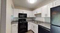 This two-story residence is designed with functionality and comfort in mind. The kitchen is well-appointed with crisp white shaker-style cabinetry accented by brushed nickel hardware, contrasting black appliances including an electric range, over-the-range microwave, and dishwasher. Gray laminate countertops and a double stainless steel sink complete the space, while durable wood-look vinyl flooring adds a clean, modern touch.

The open living area flows naturally from the kitchen, featuring the same warm wood-look flooring and neutral wall tones that lend a cohesive, inviting ambiance throughout the main level. A staircase connects the two floors, with the upper level housing the bedrooms.

The bedrooms are carpeted in soft gray tones and feature ceiling fans with integrated lighting. Each room benefits from natural light provided by windows fitted with horizontal blinds. Closet access is also present in the bedrooms, offering practical storage solutions.

The bathrooms maintain a clean, functional aesthetic. One features a white vanity with an integrated sink, a large mirror, and a tub-shower combination, while brushed nickel fixtures and towel bars provide consistent finishing details throughout. Neutral wall tones tie the bathrooms seamlessly into the overall design language of the space.