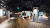 This expansive single-floor space showcases a refined industrial aesthetic, combining raw architectural elements with carefully curated furnishings. The black open-grid ceiling spans the entire floor plate, creating a dramatic overhead canopy that contrasts with warm wide-plank hardwood flooring found throughout most of the space. Exposed brick walls add texture and character, while structural columns punctuate the open layout.

A central bar serves as a focal point, featuring under-lit countertops and lined with rust-toned bar chairs on sleek metal frames, illuminated by an array of pendant and wall-mounted lights. Adjacent to the bar, a lounge area is furnished with leather sofas, accent chairs, area rugs, and a linear fireplace, fostering an inviting, relaxed atmosphere.

Toward one end, large black-framed glass walls and windows offer views of an outdoor courtyard, flooding that zone with natural light. A transitional area with herringbone brick flooring connects the interior to the exterior. Additional seating nooks with built-in banquettes and bar-height workstations provide versatile settings throughout. Glass-enclosed private rooms offer quieter, separated spaces within the larger floor plan.