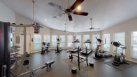 This fitness center offers a comprehensive workout environment designed to accommodate a wide range of exercise routines. The space is organized across a single floor, featuring dedicated zones for cardio, strength training, and free weights. Multiple treadmills, elliptical trainers, a recumbent bike, and an upright stationary bike make up the cardio section, while a full dumbbell rack with varied weight increments, a barbell with weight plates, and a flat weight bench serve the strength training area. A dedicated free-weight zone is enhanced by floor-to-ceiling mirrors that visually expand the space and allow users to monitor their form. The rubber-mat flooring throughout provides both safety and durability. Abundant natural light streams in through numerous windows fitted with white horizontal blinds, creating a bright and energizing atmosphere. Multiple ceiling fans help maintain airflow and comfort during workouts. Recessed lighting supplements the natural light for consistent illumination throughout the day. A wall-mounted flat-screen television adds an entertainment option for users. The textured ceilings and neutral wall tones contribute to a clean, functional aesthetic, while the Texas A&M pennant décor adds a touch of local community character to the space.