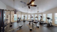 This fitness center offers a comprehensive workout environment designed to accommodate a wide range of exercise routines. The space is organized across a single floor, featuring dedicated zones for cardio, strength training, and free weights. Multiple treadmills, elliptical trainers, a recumbent bike, and an upright stationary bike make up the cardio section, while a full dumbbell rack with varied weight increments, a barbell with weight plates, and a flat weight bench serve the strength training area. A dedicated free-weight zone is enhanced by floor-to-ceiling mirrors that visually expand the space and allow users to monitor their form. The rubber-mat flooring throughout provides both safety and durability. Abundant natural light streams in through numerous windows fitted with white horizontal blinds, creating a bright and energizing atmosphere. Multiple ceiling fans help maintain airflow and comfort during workouts. Recessed lighting supplements the natural light for consistent illumination throughout the day. A wall-mounted flat-screen television adds an entertainment option for users. The textured ceilings and neutral wall tones contribute to a clean, functional aesthetic, while the Texas A&M pennant décor adds a touch of local community character to the space.