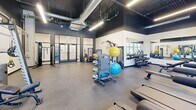 Located at 1314 Spring Garden St in Philadelphia's Spring Garden neighborhood, this fitness center delivers a well-appointed exercise experience across a single, open floor. The space is defined by its bold industrial aesthetic—dark-painted ceilings with exposed ductwork and conduit contrast sharply against crisp white walls, while large windows flood the interior with natural light and offer street-level views of the surrounding cityscape. Motivational wall graphics, including a bold "YES. YOU. CAN." mural and a gold athletic silhouette, energize the environment throughout. Durable rubber flooring covers the entire workout area, providing safety and comfort underfoot. The cardio zone features a row of treadmills and a rowing machine positioned to take advantage of the abundant window light. Strength training is well-supported with a Smith machine, cable systems, a full dumbbell rack, adjustable benches, and a wall-mounted pull-up rig. A dedicated functional training area houses medicine balls, stability balls, TRX suspension trainers, and open floor space for dynamic movement. Floor-to-ceiling mirrors expand the sense of space and assist with form. The glass-paneled entry doors add a polished, welcoming touch to this comprehensively equipped fitness facility.