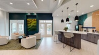 Located at 4100 Lonas Pkwy in Midlothian, Virginia, this single-story amenity clubhouse is a thoughtfully designed community hub that blends contemporary aesthetics with practical functionality. The entry foyer welcomes visitors with herringbone-patterned light wood flooring, elevated ceilings with slatted detailing, and bold teal accent walls adorned with a striking large-scale moss art installation that anchors the space with a natural, organic focal point. Tasteful furnishings, including boucle seating arrangements and warm amber leather accent chairs, create an inviting atmosphere throughout the lounge areas. A central bar-height island with a white quartz countertop serves as a communal gathering point, illuminated by a cluster of modern matte black pendant lights. The kitchen and refreshment station features a dramatic tiled backsplash in teal and terracotta hues, dark wood cabinetry, and a dedicated coffee and beverage bar. French doors along multiple walls allow natural light to pour in and provide outdoor views and access. A dedicated co-working area with individual workstations offers a focused setting for residents. The expansive fitness center is equipped with a comprehensive array of cardio machines, strength-training equipment, and free weights, all set against mirrored walls and large windows that bring in ample natural light.
