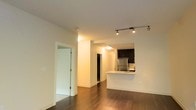 This single-floor space showcases a thoughtfully designed open-concept layout that seamlessly blends the living area with a well-appointed kitchen. The kitchen features dark cabinetry, stainless steel appliances including a refrigerator, and a clean white breakfast bar peninsula that provides both additional counter space and a casual dining option. Track lighting above the kitchen adds focused illumination, complementing the recessed lighting throughout the unit.

The flooring is a standout feature — rich, dark hardwood runs throughout the main living area, adding warmth and a polished aesthetic that contrasts beautifully with the crisp white walls and ceilings. The open hallway leads to additional rooms, and a glass-paneled door to one side introduces a modern architectural detail that allows light to pass between spaces while maintaining separation.

The overall ambiance is clean, minimalist, and contemporary, with a neutral color palette that provides a versatile canvas for any interior style. The layout is efficient yet open, creating a sense of spaciousness. The combination of quality finishes, modern lighting, and cohesive design elements makes this a well-crafted and functional single-floor space.