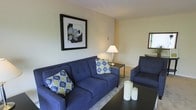 This single-floor residence offers a thoughtfully arranged living space designed for comfort and functionality. The living area serves as the heart of the home, featuring rich navy blue upholstered seating — including a full-sized sofa and coordinating armchair — accented with vibrant patterned throw pillows that add a pop of color and personality. A sleek dark coffee table anchors the seating arrangement, complemented by a classic table lamp that provides warm, ambient lighting. The walls are finished in soft, neutral tones that create a calm and inviting atmosphere, while a boldly framed botanical art piece adds a striking focal point to the room. A decorative mirror near the window reflects natural light, enhancing the sense of openness. The clean lines and understated décor throughout suggest a space that balances practicality with style. The neutral flooring and area rug tie the room together, contributing to a cohesive and polished aesthetic. Overall, this residence presents a comfortable and well-maintained living environment, ideal for those seeking a move-in-ready space with a modern yet approachable character.