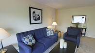 This single-floor residence offers a thoughtfully arranged living space designed for comfort and functionality. The living area serves as the heart of the home, featuring rich navy blue upholstered seating — including a full-sized sofa and coordinating armchair — accented with vibrant patterned throw pillows that add a pop of color and personality. A sleek dark coffee table anchors the seating arrangement, complemented by a classic table lamp that provides warm, ambient lighting. The walls are finished in soft, neutral tones that create a calm and inviting atmosphere, while a boldly framed botanical art piece adds a striking focal point to the room. A decorative mirror near the window reflects natural light, enhancing the sense of openness. The clean lines and understated décor throughout suggest a space that balances practicality with style. The neutral flooring and area rug tie the room together, contributing to a cohesive and polished aesthetic. Overall, this residence presents a comfortable and well-maintained living environment, ideal for those seeking a move-in-ready space with a modern yet approachable character.