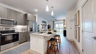 Located at 12221 Gregg Manor Rd in Manor, TX, this 1-bedroom, 1-bathroom residence offers a thoughtfully designed open-concept floor plan with a clean, contemporary aesthetic throughout. Light wood-style flooring flows seamlessly across the living areas, complementing the bright, neutral palette.

The kitchen is a standout feature, showcasing sleek gray wood-grain cabinetry that extends to the ceiling, a textured tile backsplash, stainless steel appliances — including a refrigerator, range, and microwave — and a granite-topped island with bar seating, illuminated by modern pendant lights. The kitchen opens directly to the combined dining and living area, where large sliding glass doors flood the space with natural light and provide access to a private balcony overlooking a community pool area.

The spacious bedroom features large windows with outdoor views, a ceiling fan, and durable wood-style flooring. Two closets, including a well-organized walk-in, offer generous storage. The full bathroom is finished with large-format tile flooring, a floating vanity with granite countertop, and a soaking tub with tiled surround. A dedicated laundry area adds everyday convenience. This residence offers a polished, modern living experience in a vibrant community setting.