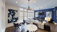 Located at Madison Square in Charlotte, NC, this single-floor community clubhouse offers a thoughtfully designed open-concept space that seamlessly blends social, dining, and lounge functions. Rich hardwood flooring runs throughout, anchoring the contemporary aesthetic defined by bold navy blue accent walls, crown molding, and an array of curated artwork.

The expansive lounge area is bathed in natural light thanks to a striking wall of large double-hung windows with transom panels above, offering direct views of the outdoor pool and surrounding greenery. Statement globe and pendant chandeliers add a stylish, modern ambiance, while plush sectional seating and small café-style tables create versatile gathering spots for residents.

The full kitchen is a standout feature, equipped with stainless steel appliances, granite countertops, white shaker cabinetry, mosaic tile backsplash, and a large central island with bar seating for several guests. Industrial-style pendant lighting illuminates the island, making it an ideal space for casual dining and entertaining. A Starbucks coffee station is also present, adding a premium amenity touch.

Throughout the space, large abstract paintings contribute to a vibrant, curated atmosphere. The overall design strikes a balance between sophisticated and welcoming, making this clubhouse a well-appointed hub for community life.