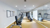 This fitness center is designed to support a well-rounded exercise routine in a clean, functional environment. The space is outfitted with a variety of cardio machines, including treadmills and an elliptical trainer, alongside a weight bench for strength training. Large floor-to-ceiling mirrors line one wall, visually expanding the room and allowing users to monitor their form during workouts. Natural light filters in through slatted window blinds, complementing the bright recessed lighting installed throughout the ceiling, which ensures the space is well-lit at any time of day. The flooring features a durable, cushioned surface in a warm neutral tone, providing comfort and support underfoot. The walls are finished in a crisp white, contributing to the bright and open atmosphere of the room. A bulletin board is mounted near the entrance, likely for posting schedules or facility guidelines. The layout is thoughtfully arranged to allow easy movement between equipment. Overall, the fitness center offers a practical and welcoming space for residents or members to maintain an active lifestyle.