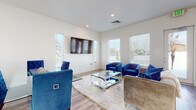 This well-appointed single-floor space combines contemporary design with functional living. The living area makes a striking visual statement with plush royal blue velvet armchairs and a coordinating sofa arranged around a sleek, chrome-accented glass coffee table atop a plush cream area rug. Light wood-tone flooring runs throughout, complementing the clean white walls and creating a bright, cohesive aesthetic. Multiple windows fitted with white horizontal blinds allow natural light to fill the room, while a wall-mounted flat-screen television serves as a focal point. Recessed ceiling lighting provides warm, even illumination throughout.

The kitchen is a standout feature, showcasing a large central island with a smooth quartz countertop and matching textured wood-grain cabinetry that extends to the walls. Stainless steel appliances — including a full-size refrigerator, dishwasher, and microwave — lend a modern, professional quality to the space. Pendant lights hang above the island, adding both task lighting and a refined design touch. The soaring ceiling height in the kitchen area enhances the sense of openness, while a window brings in natural light. The overall layout flows seamlessly between the living and kitchen areas, making this a versatile and inviting space.