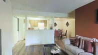 This single-floor residence features an open-concept layout that connects the living, dining, and kitchen areas in a cohesive and functional arrangement. The living area is anchored by rich hardwood flooring and a classic textured ceiling, providing a warm and inviting atmosphere. A low half-wall separates the kitchen from the main living space while maintaining an open sightline throughout. The kitchen is equipped with light wood cabinetry, white appliances including a refrigerator and range, and a countertop that doubles as a casual bar or serving surface. Recessed lighting illuminates the kitchen area effectively. The dining area, visible beyond the half-wall partition, accommodates a full dining table and chairs, complemented by decorative wall accents that add a touch of character to the space. The living room offers ample space for seating and everyday use. Neutral wall tones throughout create a clean, versatile backdrop, while the consistent hardwood flooring ties each area together seamlessly. The overall ambiance is practical and comfortable, with a layout well-suited for both everyday living and casual entertaining.