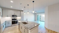 This well-appointed single-floor residence offers a thoughtfully designed open-concept layout encompassing one bedroom, two bathrooms, a kitchen, living room, and a walk-in closet. The kitchen serves as the heart of the home, showcasing a large central island with granite countertops, an undermount sink, and stylish pendant lighting. White shaker-style cabinetry extends to the ceiling, providing generous storage, while stainless steel appliances—including a refrigerator, range, microwave, and dishwasher—complete the space. Light wood-style flooring flows throughout the main living areas, creating a bright and cohesive aesthetic. The living room is airy and inviting, featuring a ceiling fan and sliding glass doors that open onto a private balcony with open outdoor views. The bedroom offers plush carpeting, a ceiling fan, and a large window that fills the room with natural light. It connects to a spacious walk-in closet with dual hanging rods and built-in shelving. The primary bathroom features granite countertops, a dual-sink vanity with white cabinetry, and tall ceilings that enhance the sense of openness. Clean white walls and modern finishes throughout lend the space a fresh, contemporary character.