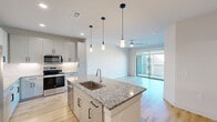 This well-appointed single-floor residence offers a thoughtfully designed open-concept layout encompassing one bedroom, two bathrooms, a kitchen, living room, and a walk-in closet. The kitchen serves as the heart of the home, showcasing a large central island with granite countertops, an undermount sink, and stylish pendant lighting. White shaker-style cabinetry extends to the ceiling, providing generous storage, while stainless steel appliances—including a refrigerator, range, microwave, and dishwasher—complete the space. Light wood-style flooring flows throughout the main living areas, creating a bright and cohesive aesthetic. The living room is airy and inviting, featuring a ceiling fan and sliding glass doors that open onto a private balcony with open outdoor views. The bedroom offers plush carpeting, a ceiling fan, and a large window that fills the room with natural light. It connects to a spacious walk-in closet with dual hanging rods and built-in shelving. The primary bathroom features granite countertops, a dual-sink vanity with white cabinetry, and tall ceilings that enhance the sense of openness. Clean white walls and modern finishes throughout lend the space a fresh, contemporary character.