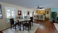 This single-floor space is defined by an open, flowing layout that connects the dining and kitchen areas into one cohesive living environment. Rich hardwood floors run throughout, providing warmth and continuity across the space.

The dining area features a high-top table paired with dark-upholstered bar-height chairs, set atop a plush cream area rug that adds texture and contrast. Framed artwork adorns the warm beige walls, and multiple large windows flood the room with natural light while offering views of the surrounding greenery outside.

A ceiling fan with a classic design is centrally positioned overhead, ensuring comfortable airflow throughout the open layout. The kitchen, visible from the dining area, boasts warm honey-toned cabinetry and a peninsula-style breakfast bar with matching bar stools — ideal for casual dining or entertaining. Pendant lights hang above the counter, providing focused task lighting that complements the overall warmth of the space.

The neutral color palette of soft whites and beiges, combined with the natural wood tones of the flooring and cabinetry, creates a welcoming and cohesive aesthetic. The open-concept design maximizes the sense of space, making this single-floor layout feel airy, functional, and well-suited for both everyday living and hosting guests.