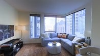 This single-floor urban residence offers a refined living experience defined by its striking corner windows that flood the interior with natural light and frame impressive city skyline views. The living area is a focal point of the space, anchored by a plush L-shaped sectional sofa in a soft blue-gray tone, accented with warm terracotta and neutral throw pillows. A sleek, round white coffee table sits atop a richly textured area rug, adding depth and warmth to the room. The wraparound floor-to-ceiling windows create a seamless connection to the vibrant cityscape beyond, showcasing a dense urban panorama of high-rise buildings and architectural detail. Roller shades offer flexible light control without compromising the expansive views. Warm ambient lighting from a tall floor lamp complements the natural light, creating a welcoming atmosphere throughout the day and into the evening. The overall design balances contemporary elegance with comfortable, livable style, making this a sophisticated urban retreat with a strong sense of place and an undeniable connection to the energy of the city surrounding it.