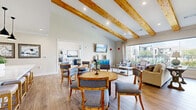 Located at 1500 Highway 90 W in Sealy, Texas, this well-appointed single-floor space offers a warm and inviting atmosphere designed for both comfort and functionality. Soaring vaulted ceilings adorned with natural wood beams lend rustic charm throughout, while light wood-tone flooring flows seamlessly across the open layout.

The living area is generously proportioned, bathed in natural light from expansive floor-to-ceiling windows that frame outdoor views of manicured landscaping and a community pool area. Comfortable seating arrangements, a wall-mounted TV, and a vibrant blue accent cabinet create an inviting, lived-in feel. A second seating area offers additional space for relaxation, complete with plaid armchairs and a large coffee table.

The combined dining and kitchen area is a standout feature of the space. Multiple round dining tables with upholstered chairs provide ample seating, while the kitchen impresses with its crisp white cabinetry, subway tile backsplash, open shelving, stainless steel appliances, and a large central island topped with white quartz and accented by sleek black pendant lights. A shuffleboard table and additional seating nook round out the space, making it ideal for social gatherings.