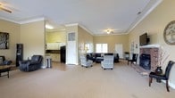 This single-floor space offers an open and functional layout designed for comfort and versatility. The main area is generously proportioned, featuring warm beige walls accented by crisp white crown molding that adds a refined architectural touch throughout. A ceiling fan overhead and multiple windows allow for ample natural light, creating a bright and welcoming ambiance.

The lounge area is furnished with a large sectional sofa and a pair of patterned accent chairs, arranged to encourage comfortable conversation and gathering. The carpeted flooring adds warmth and softness underfoot, while the open floor plan provides flexibility for various uses and configurations.

Adjacent to the main lounge, a kitchenette area is visible and includes white cabinetry, a dark refrigerator, and a countertop workspace. This functional nook is well-suited for light food and beverage preparation, making the space ideal for events or community gatherings.

Additional doors and passageways suggest further utility rooms or access points, contributing to the overall practicality of the layout. The combination of comfortable furnishings, thoughtful architectural detailing, and a well-integrated kitchenette makes this a versatile and inviting space suited for a wide range of social or organizational purposes.