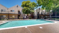 Located at 7200 Gloria Dr in Sacramento, California, this outdoor pool area serves as a central communal amenity within a multi-building residential complex. The generously sized rectangular swimming pool is the focal point of the space, featuring clear turquoise water, in-pool lighting, and entry steps with a metal handrail for easy access. Surrounding the pool is a spacious concrete and aggregate deck finished in a warm terracotta tone, providing ample room for lounge chairs and outdoor seating arrangements. A poolside information and safety station is conveniently positioned near the water's edge, complete with posted guidelines and a life ring. The area is enclosed by a black wrought iron fence with gated access points, ensuring both security and visibility. Mature deciduous trees provide generous natural shade across portions of the deck, enhancing comfort during Sacramento's warm sunny days under open blue skies. The surrounding two-story stucco buildings feature distinctive gabled rooflines with circular accent windows, covered balconies, and exterior staircases, contributing to a cohesive architectural character. The well-maintained landscaping with shrubs and greenery adds to the inviting outdoor atmosphere of this community gathering space.