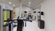 The Warehouse Fitness Center in Somerville, NJ offers a well-organized, single-floor fitness facility designed to accommodate a variety of workout needs. The main exercise area features soaring vaulted ceilings that lend an open, airy atmosphere, complemented by recessed lighting throughout. The floors are finished with a combination of light-colored hard flooring and yellow rubber matting in the primary workout zone, providing both durability and safety during exercise.

The facility is equipped with a range of strength and cardio machines, including a multi-station cable machine, weight benches, treadmills, and additional resistance equipment. Large wall-mounted mirrors are strategically positioned to allow users to monitor their form during workouts. A wall-mounted flat-screen television and a dedicated wall-mounted air conditioning unit add to the comfort and convenience of the space.

A separate enclosed room, accessible through a glass-paneled sliding door, provides a more private area for cardio training, housing a treadmill and additional equipment. A water cooler station is conveniently available within the main area. The neutral, light-toned walls and clean finishes throughout contribute to a modern, professional atmosphere suited for focused, effective training sessions.