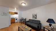 This single-floor unit offers a practical and comfortable layout designed to maximize every square foot. The open-concept living and dining area is anchored by warm hardwood flooring and neutral white walls, creating a bright and welcoming atmosphere. A tufted gray sofa, rustic wood coffee table, and urban-themed wall art give the living space a modern yet homey character, while a compact dining set for two sits conveniently adjacent to the kitchen.

The kitchen is efficiently designed with warm-toned wood cabinetry, dark granite-style countertops, a stainless steel sink, and white appliances including a gas range and full-size refrigerator. A range hood and overhead cabinets provide ample storage and ventilation.

The bedroom is a calm retreat with soft gray walls, rich hardwood floors, and a queen-size bed framed by a dark wood headboard. A dedicated closet with a hanging rod offers organized storage space.

The bathroom features clean white walls, light ceramic tile flooring, a wood vanity with countertop sink, and a tub-shower combination enclosed by a decorative patterned curtain. Flush-mount ceiling lighting throughout the unit ensures consistent illumination across all areas.