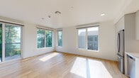 This single-floor unit presents a cohesive, modern aesthetic defined by crisp white walls and warm light wood flooring that runs continuously throughout the space. The open-concept kitchen is a standout feature, equipped with flat-front gray cabinetry extending floor to ceiling, stainless steel appliances including a range, dishwasher, and refrigerator, complemented by a white subway tile backsplash and light-toned countertops. Recessed lighting illuminates the kitchen area effectively. The living area adjoins the kitchen, offering an open and versatile layout with clean sightlines. Additional rooms provide flexible use, with at least one bedroom benefiting from a large window overlooking a lush, green outdoor setting, allowing natural light and garden views. The bathroom features a navy-toned vanity with ample storage, a white countertop, and a large mirror. A dedicated in-unit laundry area with a stacked washer and dryer is conveniently located nearby. Throughout the unit, white baseboards, minimalist hardware, and a neutral palette contribute to a clean, contemporary atmosphere.