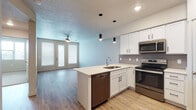 This well-appointed single-floor residence showcases a thoughtfully designed open-concept layout that seamlessly connects the kitchen and living areas. The kitchen is a standout feature, boasting crisp white cabinetry, a gray tile backsplash, and a full suite of stainless steel appliances including a refrigerator, electric range, over-the-range microwave, and dishwasher. A peninsula island with a white countertop and integrated sink provides additional workspace and casual seating, while stylish cylindrical pendant lights hang overhead, adding a contemporary touch. Light wood-style flooring runs throughout the main living areas, enhancing the bright and airy atmosphere. The open living space extends toward glass-paneled doors that invite natural light and offer access to the exterior. A ceiling fan serves both the living area and adjoining spaces. The bedroom features plush neutral carpeting and a clean, minimalist design, offering a comfortable and quiet retreat. The bathroom is finished with a clean white tub-shower combination with built-in corner shelving and continues the light wood-style flooring found throughout the unit. Textured ceilings add a classic touch, while the light blue-toned walls create a calm, cohesive palette across the space.