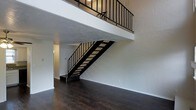 This two-story space makes a strong first impression with its open, airy layout and clean, modern aesthetic. The main floor is anchored by rich, dark hardwood flooring that contrasts beautifully against the bright white walls, creating a sleek and contemporary atmosphere throughout. A standout architectural feature is the bold, industrial-style staircase — finished in matte black metal with open wooden treads — that rises to the upper level. The staircase is bordered by a matching black metal railing that continues along the second-floor mezzanine, offering an open view down to the main living area below. The upper level overlooks the main floor through this open railing system, enhancing the sense of vertical space and connectivity between floors. The generous ceiling height amplifies the feeling of openness and volume, making the interior feel expansive and unenclosed. The neutral white walls throughout both floors provide a versatile backdrop suitable for a variety of uses and décor styles. Additional doorways visible on the main level hint at further rooms beyond the open living area, offering practical functionality alongside the dramatic design elements. Overall, the space combines industrial character with a clean, minimalist sensibility.