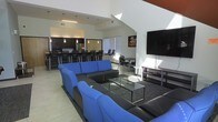 This two-story space is designed as a versatile communal gathering area, combining multiple functional zones within an open, airy layout. The soaring ceilings create an impressive sense of volume, drawing the eye upward and enhancing the overall spaciousness of the interior.

The living area is anchored by a large sectional sofa in bold blue and black upholstery, arranged around a glass-top coffee table atop a area rug — creating a comfortable lounge setting ideal for socializing. A wall-mounted flat-screen television is positioned prominently, accompanied by a media console, making the space well-suited for entertainment.

Adjacent to the lounge, a dining zone features compact tables and task-style chairs, offering a more structured seating arrangement. The kitchen area, visible in the background, is outfitted with dark cabinetry, stainless steel appliances, and a long bar-height counter lined with matching black barstools — providing ample seating for casual dining or social gatherings.

Natural light filters through horizontal-blind-covered windows, brightening the neutral-toned walls and light tile flooring. Pendant lighting over the kitchen adds a finishing touch to this well-appointed, multi-use space.