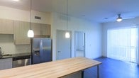This single-floor space features an open-concept design that seamlessly connects the kitchen and living areas. The kitchen is equipped with a stainless steel French door refrigerator with an ice and water dispenser, upper cabinetry in a warm wood finish, and a generous butcher block island that serves as both a prep surface and casual dining area. Two pendant lights hang above, providing focused task lighting that complements the overall brightness of the space.

The flooring transitions from what appears to be tile or light-colored material in the kitchen zone to dark hardwood or laminate in the living area, adding visual contrast and definition between the two spaces. White walls and a vaulted or elevated ceiling contribute to an airy, open atmosphere throughout.

Two interior doorways are visible, suggesting access to additional rooms such as bedrooms or a bathroom. Natural light filters in through a window on the far wall of the living area, enhancing the bright and clean aesthetic. HVAC vents are integrated into both the ceiling and upper wall areas, ensuring efficient climate control. Overall, the space offers a functional, modern layout with a neutral palette suitable for a variety of interior styles.
