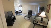 This single-floor residence features an open-concept design that seamlessly connects the living and dining areas into one cohesive, functional space. The living area is carpeted in a neutral beige tone, providing a warm and comfortable foundation throughout. Large vertical blinds on a wide window allow generous natural light to fill the room, creating a bright and airy atmosphere.

The living area is well-proportioned, accommodating a sectional sofa, an accent armchair, and a dark-finished rectangular coffee table, making it suitable for both relaxing and entertaining. A media console along the wall provides practical storage and display space.

The adjacent dining area flows naturally from the living space, featuring a round dark-wood dining table paired with matching dark upholstered chairs, ideal for everyday dining. A ceiling fan with a light fixture is centrally positioned overhead, contributing to both ventilation and ambient lighting.

The walls are finished in a soft cream tone, complementing the neutral carpet and dark furniture accents throughout. The overall ambiance of the space is casual and practical, with an emphasis on open, easy-to-navigate floor space.