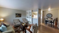 This single-floor residence offers a thoughtfully arranged layout that maximizes both comfort and functionality. The living area serves as the heart of the space, featuring neutral gray walls, tile flooring, and a coordinated seating arrangement that includes patterned accent chairs and a cushioned ottoman. A decorative area rug anchors the seating zone, adding warmth and texture to the room. A standout architectural element is the built-in open shelving unit that acts as a partial room divider, elegantly separating the living area from the kitchen while maintaining an open, airy feel. The shelving displays decorative accents and serves as both a functional and aesthetic feature. The kitchen is visible through the pass-through opening above a countertop bar, revealing white cabinetry and clean, bright finishes that contribute to the light and welcoming atmosphere. Adjacent to the kitchen, a dining area with bar-height seating provides a casual space for meals. The overall ambiance is bright and cohesive, with a neutral color palette punctuated by colorful decorative touches, including wall art and potted flowers, creating a lived-in yet polished environment throughout.