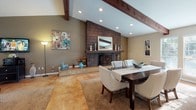 This single-story space is defined by its open-concept layout and striking architectural details. Vaulted ceilings with prominent dark wood exposed beams run throughout the main living areas, creating a sense of generous vertical space and rustic character. Polished stained concrete floors in warm amber tones flow continuously across the interior, unifying each area cohesively.

The living room is expansive and light-filled, benefiting from large sliding glass doors that provide direct outdoor views and access while allowing natural light to permeate the space. Recessed lighting supplements the natural light throughout.

A focal point of the adjoining dining area is the floor-to-ceiling tiled fireplace, clad in the same dark brown shingle-style tile that also accents the kitchen's pass-through bar. The fireplace features a wood mantel and anchors the space with warmth and visual interest.

The kitchen is efficiently arranged with dark cabinetry, black appliances, and a pass-through breakfast bar with brick-tile cladding and seating for two. A dedicated workspace area adjacent to the kitchen offers a functional office setup, bathed in natural light from nearby windows. The overall ambiance is warm, casual, and well-connected throughout.